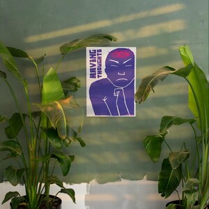 May include: A poster with the text "HAVING THOUGHTS" and a purple illustration of a person with a red brain. The person has a stern expression. The poster is mounted on a green wall, surrounded by green plants.
