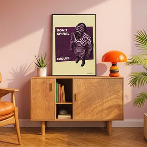 May include: A framed art print with the words "DON'T SPIRAL EVOLVE" and a graphic of a figure with zebra-like stripes. The print is displayed on a wooden cabinet with an orange mushroom lamp and a potted plant.