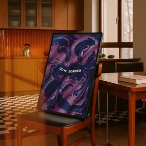 May include: A framed art print with a swirling abstract design in shades of purple and navy blue. The text "DO IT SCARED" is displayed in white. The print is propped on a wooden chair in a room with a checkered floor.