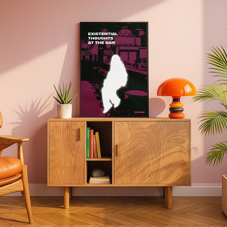 Op de afbeelding: Een ingelijste kunstdruk met de tekst "EXISTENTIAL THOUGHTS AT THE BAR" toont een wit silhouet tegen een paarse en zwarte barsc&egrave;ne. Een oranje en zwarte lamp staat op een houten kast met een plant en boeken.