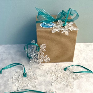 A photo of the packaging the snowflakes will come in. A recycled and recyclable kraft gift box with sparkly aqua ribbon and my logo sticker. Surrounded by 4 of the 5 snowflakes the box will contain.