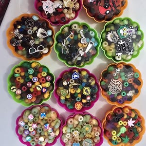 May include: Twelve colorful resin filled flower shaped containers with various charms and beads inside. The charms include a clock, a pair of sunglasses, a saw, an axe, a spider web, a skull, a heart, a flower, a planet, a marijuana leaf, a unicorn, a Pokemon character, and a Deadpool character.