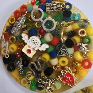 May include: A round, yellow resin coaster with a variety of charms and beads embedded in it. The charms include a pair of glasses, a feather, a snake, a frog, a Hogwarts crest, a white owl, and a lightning bolt. The beads are in various colors, including red, green, blue, yellow, and white.