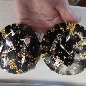 May include: Two round, black and gold resin coasters. Each coaster is embedded with small silver and gold metal tools, nuts, and bolts. The coasters are held in a person's hand.