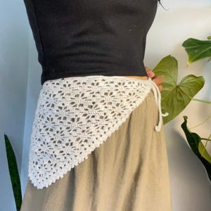 May include: A white triangular crochet wrap featuring an intricate floral-inspired openwork pattern. The garment is tied at the side with thin matching crochet strings, layered over a black short-sleeved top and a solid tan skirt.