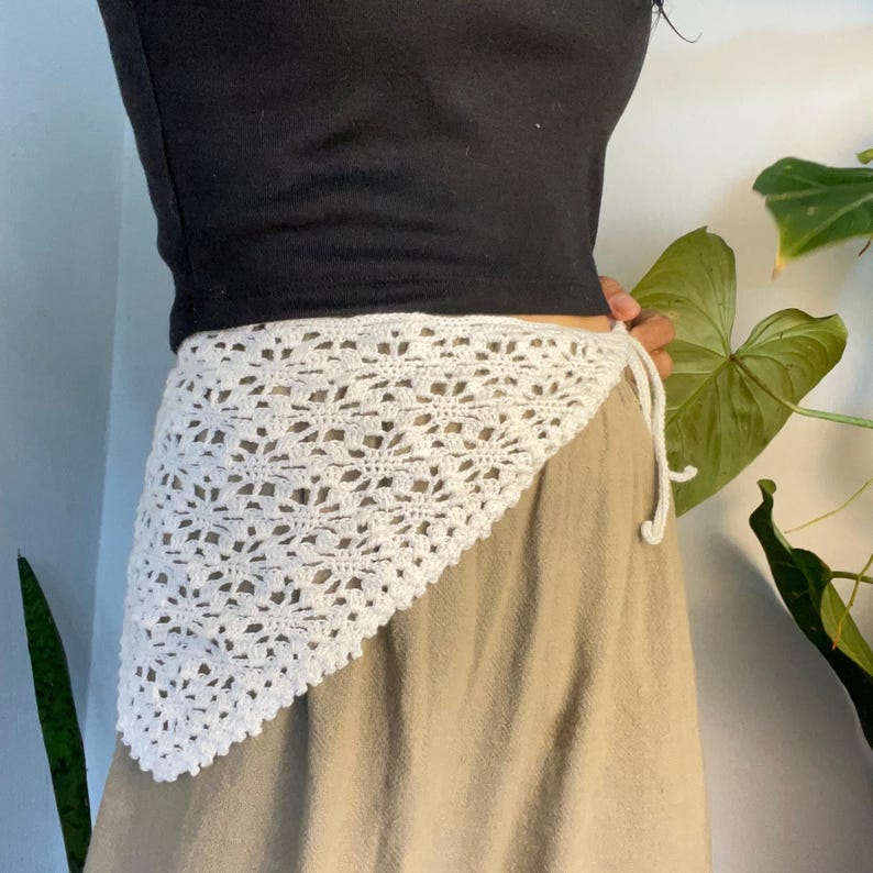 May include: A white triangular crochet wrap featuring an intricate floral-inspired openwork pattern. The garment is tied at the side with thin matching crochet strings, layered over a black short-sleeved top and a solid tan skirt.
