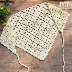 May include: Cream-colored, open-weave crochet shawl with a triangular shape. The shawl has a scalloped edge and long ties. The word "PDF" is visible in the upper right corner. The shawl is displayed on a wooden surface.