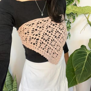 May include: A light pink triangular crochet bandana top with a repeating floral pattern worn over a black long-sleeved shirt and paired with a white skirt. The crochet piece features an intricate openwork design with scalloped edges.