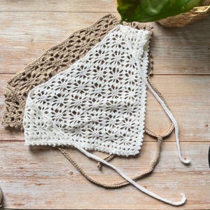 May include: Two triangular crochet headscarves, one in white and one in beige, featuring an intricate floral-inspired openwork pattern. Each headscarf includes long, thin crocheted ties at the corners for securing the garment in place.