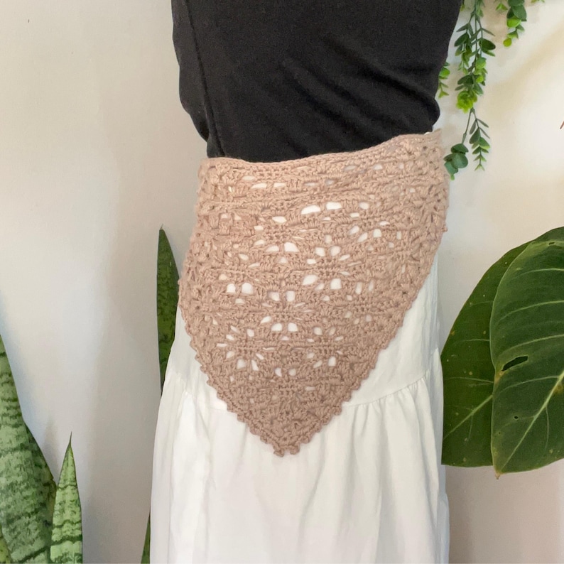 May include: A triangular crochet bandana in a light tan color featuring an intricate openwork lace pattern with a scalloped edge. The accessory is worn over a black top and a white skirt, showcasing the detailed yarn texture and craftsmanship.
