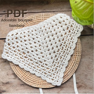 May include: A white, crocheted bandana with a scalloped edge, resting on a woven, circular placemat. The text "PDF Adorable bouquet bandana" is visible in the upper left corner. The bandana is made of a soft, open weave.