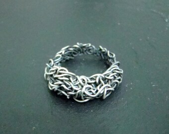 Könnte beinhalten: Ein silberner Ring aus ineinander verschlungenem Metalldraht, der ein strukturiertes, organisches Design erzeugt.