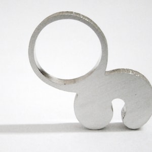 May include: A silver ring with a unique design featuring a circle and a curved shape. The ring is made of metal and has a smooth, polished finish.