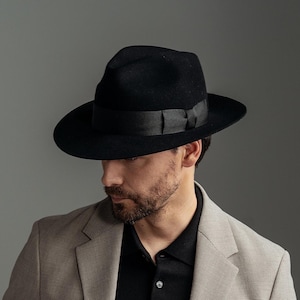 May include: A man wearing a beige suit jacket and matching trousers with a black polo shirt underneath. He is wearing a black fedora hat with a black ribbon.