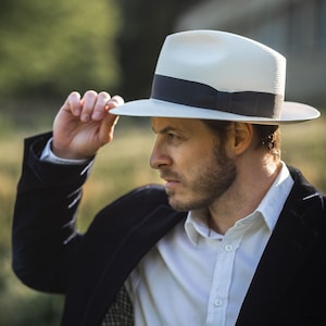 K&ouml;nnte beinhalten: Ein Mann tr&auml;gt einen wei&szlig;en Fedora mit schwarzem Band, ein wei&szlig;es Hemd mit Kn&ouml;pfen und einen schwarzen Blazer.