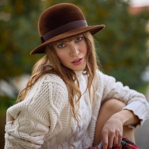 Puede incluir: Una mujer con un sombrero fedora de fieltro marrón con una banda marrón y un suéter de punto blanco. Está sentada con las piernas cruzadas y las manos en el regazo.