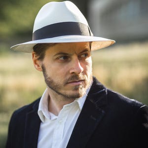 K&ouml;nnte beinhalten: Ein Mann tr&auml;gt einen wei&szlig;en Fedora mit schwarzem Band und einen schwarzen Blazer &uuml;ber einem wei&szlig;en Hemd mit Kn&ouml;pfen.