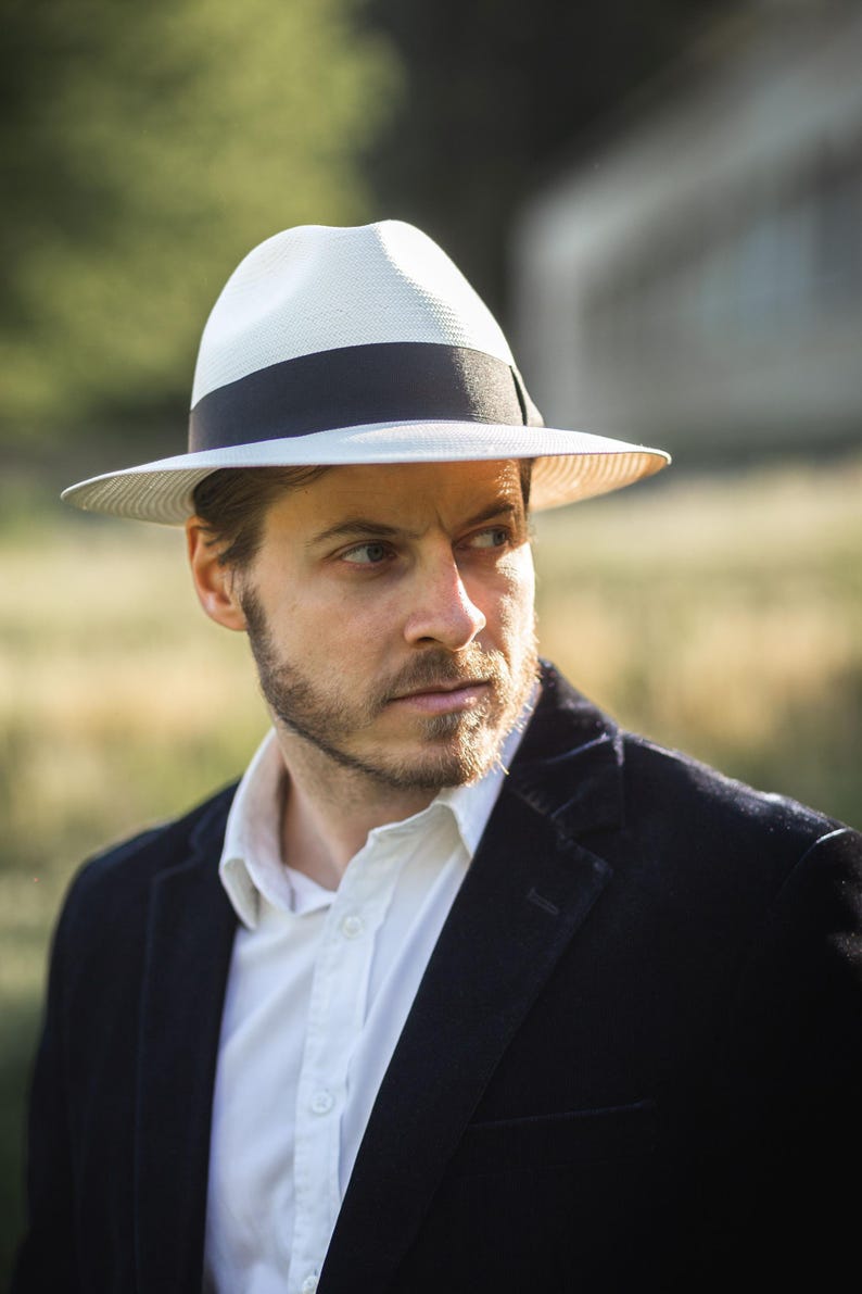 K&ouml;nnte beinhalten: Ein Mann tr&auml;gt einen wei&szlig;en Fedora mit schwarzem Band und einen schwarzen Blazer &uuml;ber einem wei&szlig;en Hemd mit Kn&ouml;pfen.