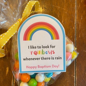 May include: A clear plastic bag filled with colorful gumballs tied with a yellow ribbon. A rainbow-themed card with the text "I like to look for rainbows whenever there is rain. Happy Baptism Day!" is attached to the bag.