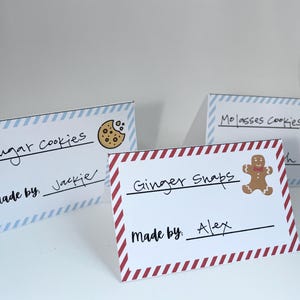 Pode incluir: Três cartões brancos com texto manuscrito e ilustrações. Um cartão diz "Sugar Cookies" com um gráfico de biscoito, outro diz "Ginger Snaps" com um boneco de gengibre e o último diz "Molasses Cookies" com um floco de neve.