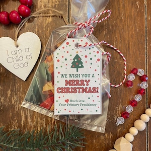 May include: A Christmas gift tag with the message "We Wish You a Merry Christmas!" and a Christmas tree graphic. The tag is tied with red and white twine to a clear cellophane bag. Other decorations include a wooden heart and red beads.