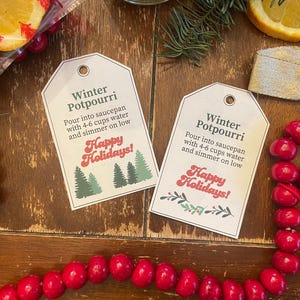 Könnte beinhalten: Zwei weiße Etiketten mit dem Text "Winter Potpourri" und "Happy Holidays!" werden auf einer Holzoberfläche angezeigt. Die Etiketten enthalten Anweisungen und Illustrationen von Bäumen. Rote Beeren und eine Orangenscheibe sind ebenfalls sichtbar.