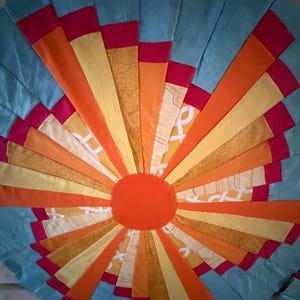 May include: A circular fabric wall hanging featuring a sunburst pattern of radiating strips in shades of light blue, bright orange, yellow, and magenta. A solid orange circle sits at the center, surrounded by alternating patterned and solid fabric segments.