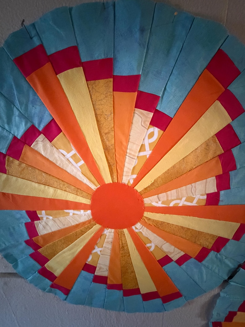 May include: A circular fabric wall hanging featuring a sunburst pattern of radiating strips in shades of light blue, bright orange, yellow, and magenta. A solid orange circle sits at the center, surrounded by alternating patterned and solid fabric segments.