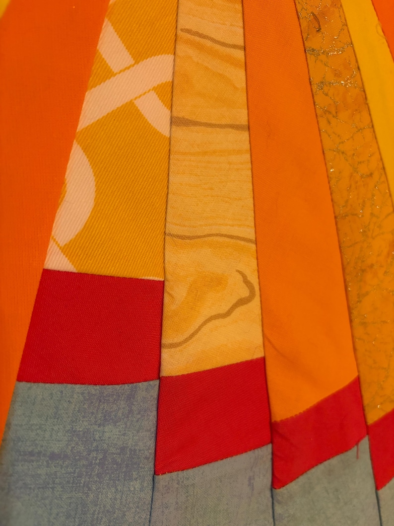 May include: A quilt featuring a radial pattern of fabric strips in shades of orange, yellow, and red, interspersed with light blue sections. The fabric textures include solid colors, a white geometric print, a wood grain pattern, and gold glitter.