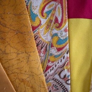 May include: A collection of fabric strips featuring a mustard yellow panel with a gold crackle pattern, a central panel with a multi-colored paisley print in red, teal, and yellow, and a bright yellow panel topped with a solid magenta horizontal band.