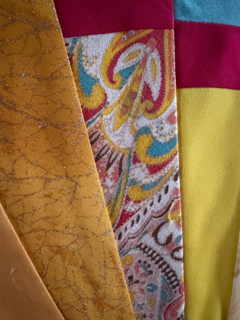 May include: A collection of fabric strips featuring a mustard yellow panel with a gold crackle pattern, a central panel with a multi-colored paisley print in red, teal, and yellow, and a bright yellow panel topped with a solid magenta horizontal band.