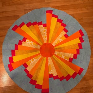 May include: A circular fabric quilt block featuring a central orange circle surrounded by radiating segments in shades of yellow, orange, and red. The outer border consists of light blue fabric panels arranged in a sunburst pattern design.