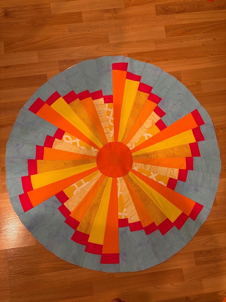 May include: A circular fabric quilt block featuring a central orange circle surrounded by radiating segments in shades of yellow, orange, and red. The outer border consists of light blue fabric panels arranged in a sunburst pattern design.