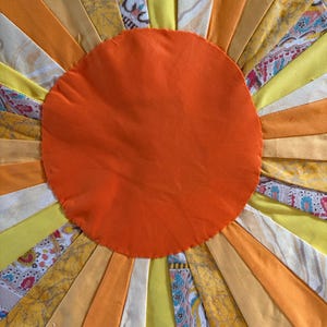 May include: A textile sunburst design featuring a solid orange circular center surrounded by radiating fabric strips in shades of yellow, orange, and cream. The strips include solid colors and various patterns, including floral and abstract prints.