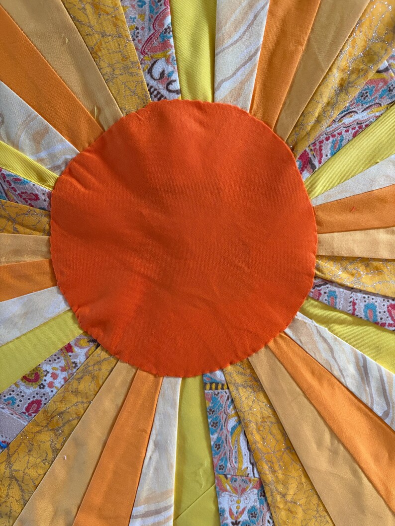 May include: A textile sunburst design featuring a solid orange circular center surrounded by radiating fabric strips in shades of yellow, orange, and cream. The strips include solid colors and various patterns, including floral and abstract prints.