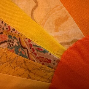 May include: A textile piece featuring a fan-shaped arrangement of fabric strips in shades of yellow, orange, and gold. The design includes solid-colored fabric, a floral print with red and green accents, and a textured gold fabric with a metallic sheen.