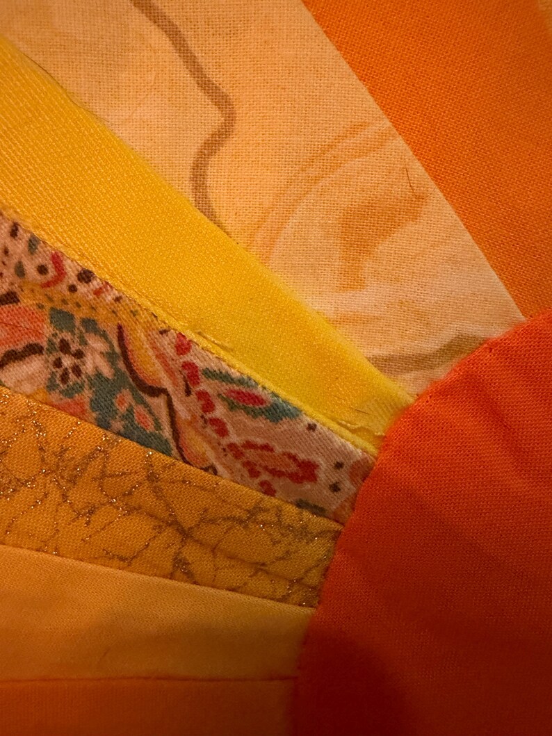 May include: A textile piece featuring a fan-shaped arrangement of fabric strips in shades of yellow, orange, and gold. The design includes solid-colored fabric, a floral print with red and green accents, and a textured gold fabric with a metallic sheen.
