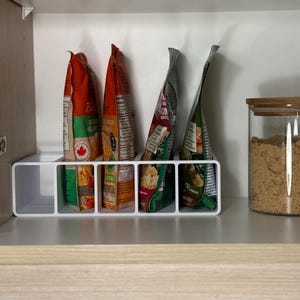 Puede incluir: Un organizador de plástico blanco dentro de un armario de madera contiene varios paquetes de alimentos. Los paquetes son de varios colores, incluyendo naranja, verde y rojo. A la derecha se ve un tarro de cristal con tapa de madera, lleno de gránulos marrones.