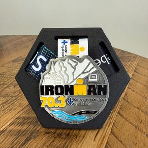 Peut inclure: Une médaille de finisseur Ironman 70.3 Mont-Tremblant Québec en métal argenté avec un motif de montagne, le texte "IRONMAN 70.3" et une vague bleue, présentée dans un support hexagonal en mousse noire avec trois rubans en tissu.