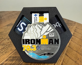 May include: A silver-toned Ironman 70.3 Mont-Tremblant Quebec finisher medal featuring a mountain graphic, the text "IRONMAN 70.3", and a blue water wave design, displayed within a black hexagonal foam holder with three fabric ribbons.