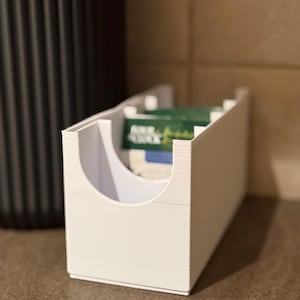 Peut inclure: Un organiseur de sachets de thé blanc et rectangulaire avec une ouverture incurvée. L'organisateur est rempli de sachets de thé et repose sur une surface marron. Un récipient noir texturé est visible en arrière-plan.