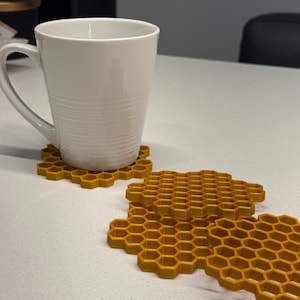 Peut inclure: Une tasse en céramique blanche repose sur un dessous de verre jaune en forme de nid d'abeille. Plusieurs autres dessous de verre sont disposés à proximité. Les dessous de verre sont en matériau texturé et présentent un motif hexagonal répétitif. La tasse et les dessous de verre sont sur une surface blanche.