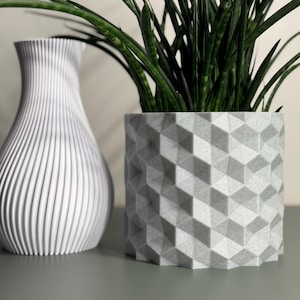 May include: A white, ribbed vase and a geometric patterned planter holding a green plant. The vase has a slender, curved shape. The planter is a light gray color and has a repeating cube design.