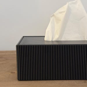 May include: A black tissue box with a ribbed design. White tissues are pulled from the top opening. The box is on a wooden surface.