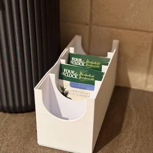 Peut inclure: Un organisateur de sachets de thé en plastique blanc avec une base rectangulaire et des découpes incurvées sur les côtés. Le contenant contient plusieurs sachets de thé verts et blancs étiquetés Four O'Clock Herbalist, Herbal Tea Calm et Good Night Bon Sommeil.