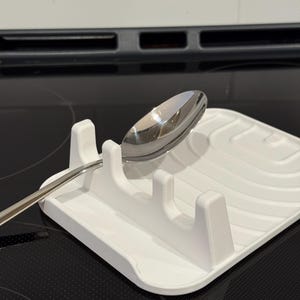 Peut inclure: Un repose-cuillère blanc avec un motif en relief et trois supports verticaux. Une cuillère en acier inoxydable repose sur le support. Le repose-cuillère est sur une plaque de cuisson noire.