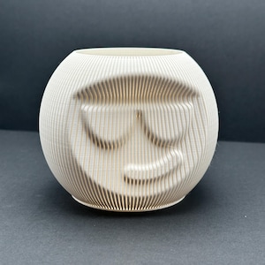 Peut inclure: Un pot décoratif sphérique blanc avec une surface texturée et nervurée. Le pot présente un visage souriant avec un design de lunettes de soleil. Le design est sculpté dans la surface, créant un effet 3D. Le pot convient aux petites plantes ou comme objet décoratif.