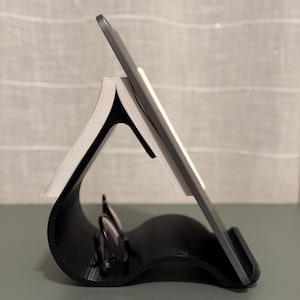 Peut inclure: Un support noir et incurvé pour livre et tablette. Le support maintient un livre et une tablette inclinés. Le support a un design ondulé et est fait d'un matériau sombre et solide. Une paire de lunettes repose dans la courbe du support.