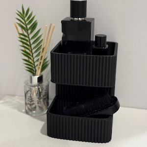 May include: A matte black tiered organiser with a vertical ribbed texture. The top tier holds a large rectangular black bottle and a smaller black bottle, while the lower tier contains a black oval brush. A glass reed diffuser sits nearby.