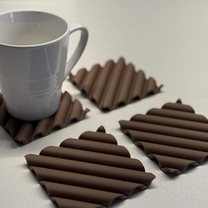Peut inclure: Une tasse en céramique blanche repose sur un dessous de verre marron et strié. Trois autres dessous de verre bruns sont dispersés à proximité. Les dessous de verre ont un design géométrique unique et sont en matériau souple.